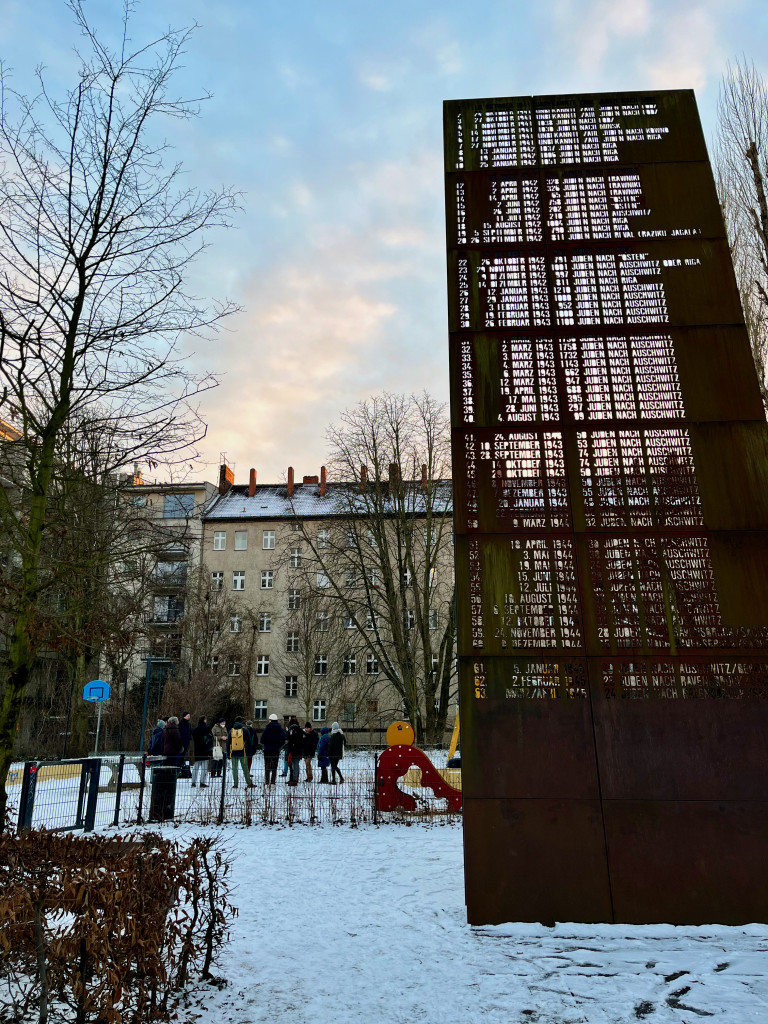 Blick auf das Mahnmal Synagoge Levetzowstraße an dem eine Gruppe steht durch eine Stele mit den eingestanzten Zügen die von dort deportiert wurden